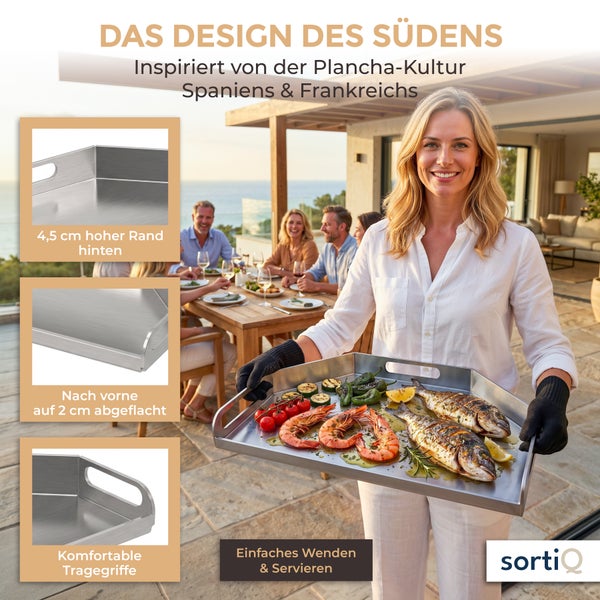 Eine Frau hält eine Edelstahl Grillplatte mit gegrilltem Fisch und Gemüse auf einer Terrasse vor einer Gruppe von Menschen.