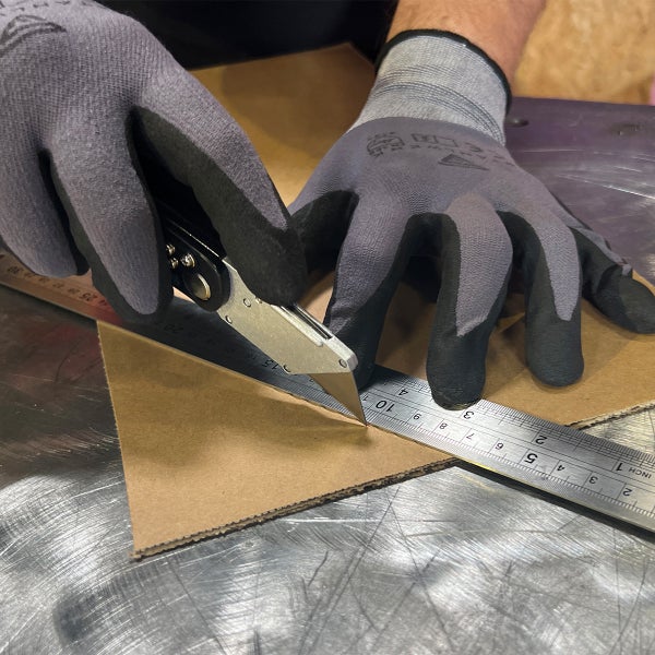 Eine Person mit Arbeitshandschuhen schneidet Karton mit einem Cuttermesser entlang eines Metalllineals auf einer Arbeitsfläche.