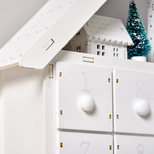 Adventskalender aus weißem Holz in Hausform mit nummerierten Schubladen und dekorativem Tannenbaum.