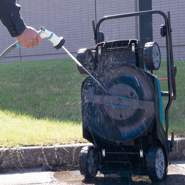 Eine Person reinigt die Unterseite eines Rasenmähers mit einem Wasserstrahl aus einer Gartenbrause.