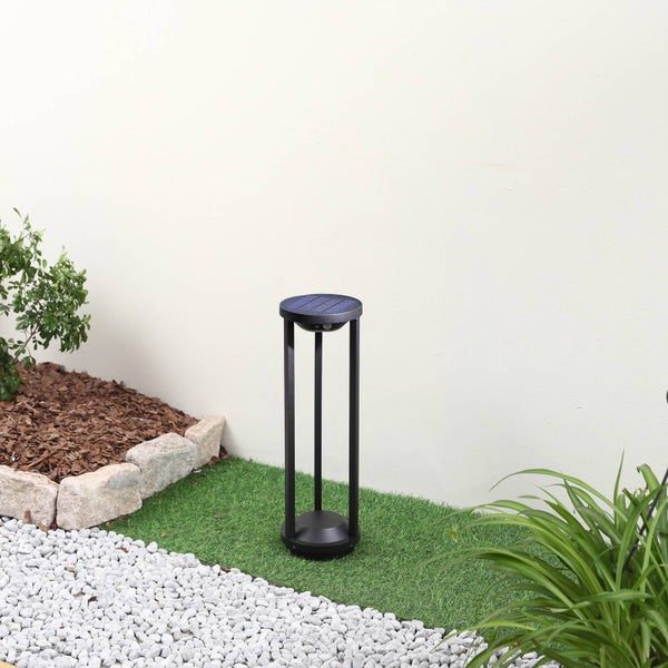 Moderne schwarze Solar-Wegeleuchte mit Solarpanel auf Kunstrasen in einer Gartenanlage mit Steinen, Mulch und Pflanzen.