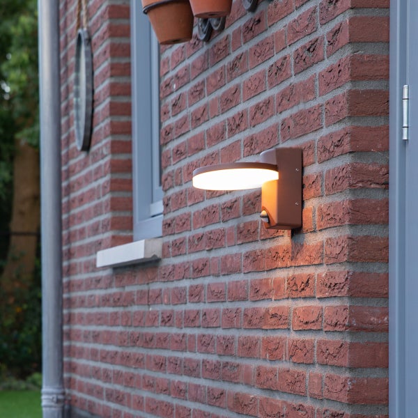 Moderne runde LED-Außenwandleuchte mit Bewegungsmelder an einer Backsteinwand montiert und eingeschaltet.