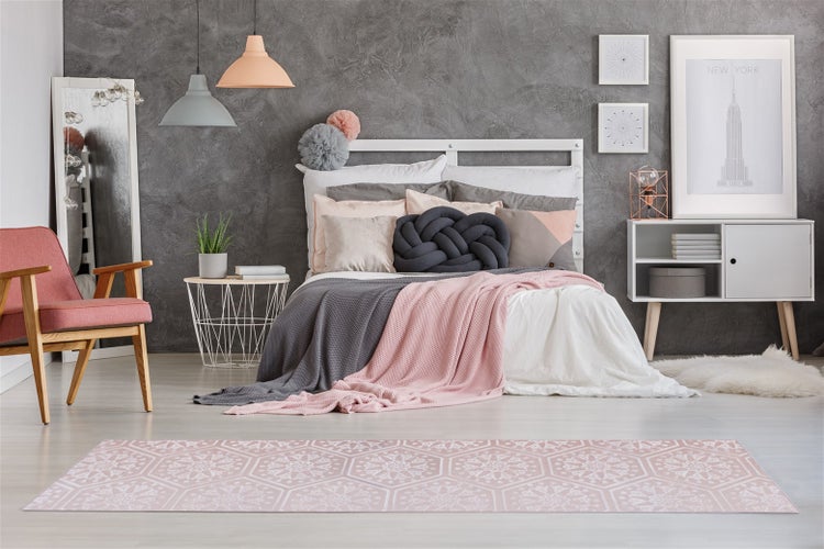 Modernes Schlafzimmer mit gemustertem Teppichläufer im Vordergrund, Doppelbett, Nachttisch, Sessel und Wanddekoration vor einer grauen Betonwand.