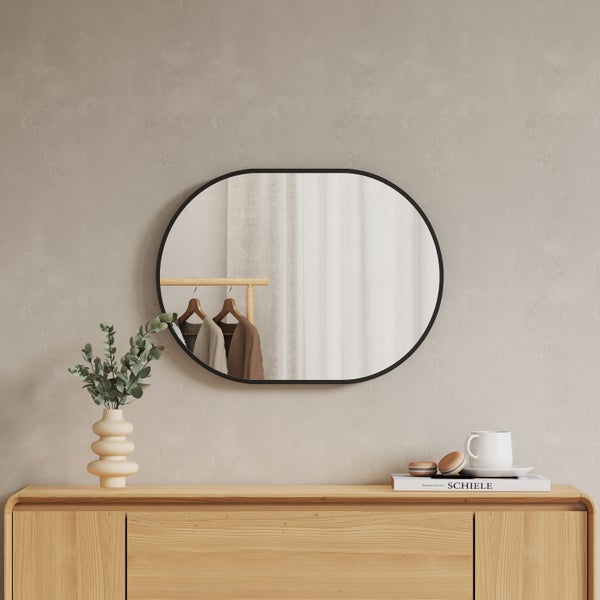 Moderner ovaler Wandspiegel mit schwarzem Metallrahmen über einem hölzernen Sideboard in einem hellen, aufgeräumten Raum.