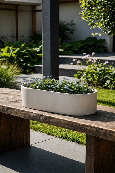 Ovale Pflanzschale aus Stein auf einer rustikalen Holzbank im sonnigen Garten mit blauen Blumen.
