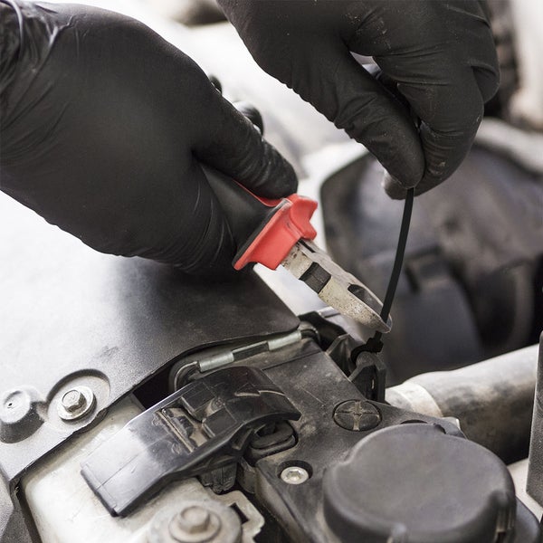 Hände in schwarzen Handschuhen schneiden mit einem Seitenschneider einen schwarzen Kabelbinder im Motorraum eines Autos durch.