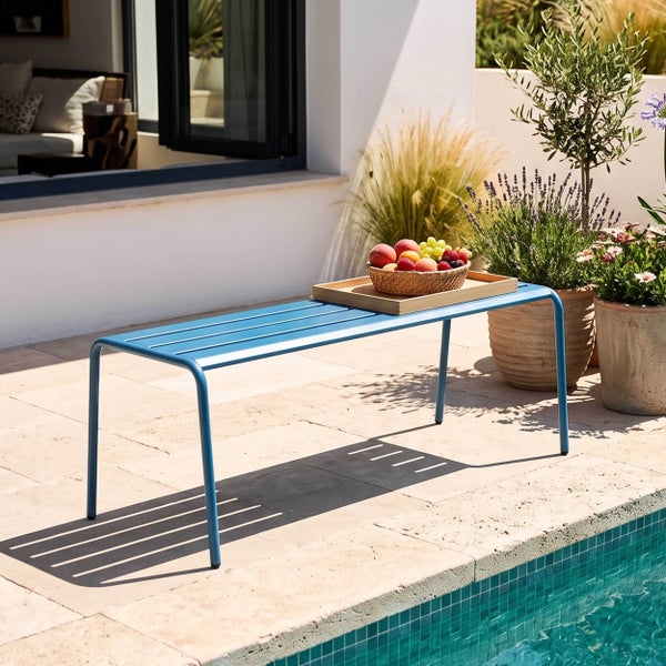 Moderne blaue Metall-Gartenbank auf einer sonnigen Steinterrasse am Pool, mit einem Holztablett mit Obst und dekorativen Topfpflanzen.