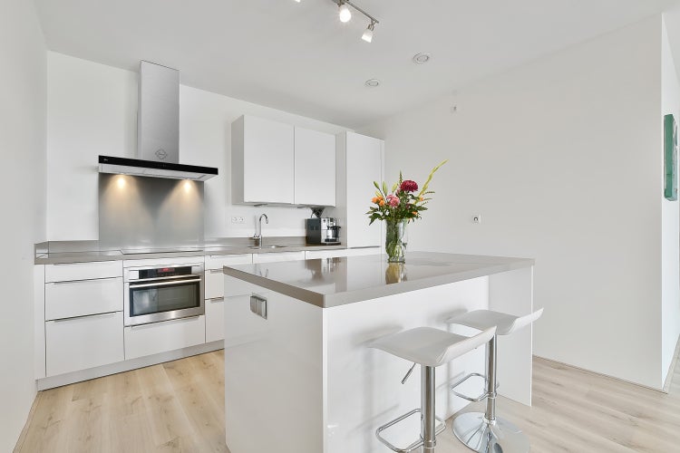 Moderne, helle Küche mit weißer Kücheninsel, grauer Arbeitsplatte, zwei weißen Barhockern, integriertem Backofen und Dunstabzugshaube aus Edelstahl.