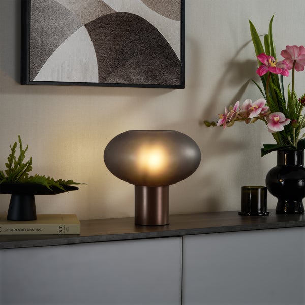 Moderne Tischleuchte in Pilzform mit ovalem Glasschirm und Metallfuß auf einer Kommode mit Dekoration und Wandbild.