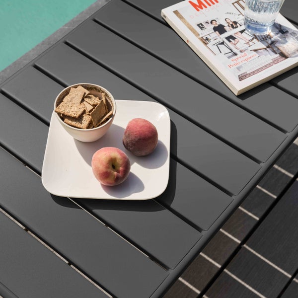 Grauer Metall-Gartentisch mit Lamellen-Tischplatte, dekoriert mit Pfirsichen, Crackern, Zeitschrift und Wasserglas an einem Swimmingpool.