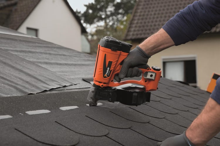 Handwerker mit Arbeitshandschuhen verwendet Paslode Druckluftnagler zur Montage von Dachschindeln auf einer Dachfläche bei einem Wohnhaus.
