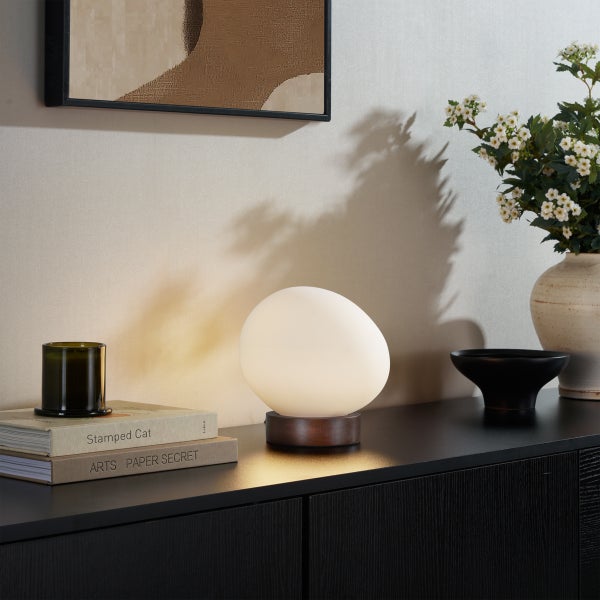 Moderne Tischlampe mit eiförmigem Leuchtmittel auf dunklem Holzsockel auf einem schwarzen Sideboard in einem stilvoll eingerichteten Wohnbereich.