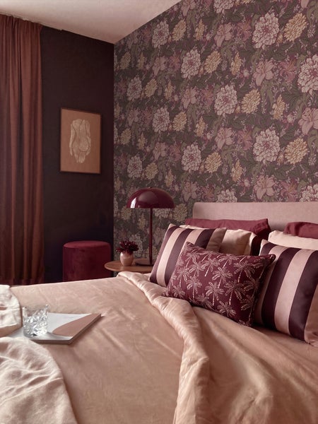 Schlafzimmer mit Vliestapete mit Blumenmuster, dunkler Wand, Bett mit rosa Bettwäsche, gemusterten Kissen und einer Tischleuchte.