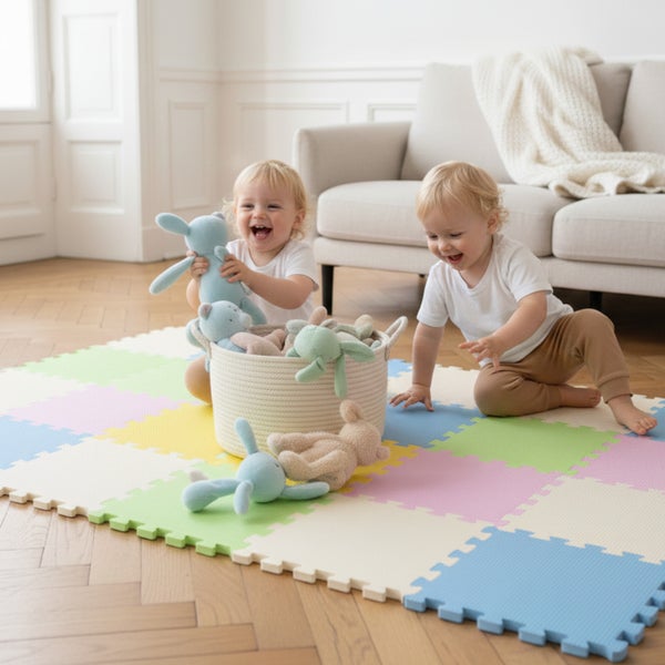 Zwei Kleinkinder spielen mit Stofftieren auf einer bunten Schaumstoff-Puzzlematte in einem hellen Wohnzimmer mit Holzboden.