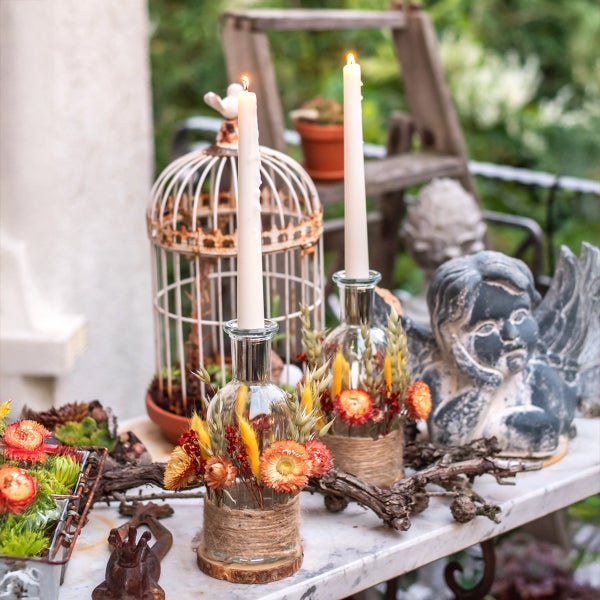 Dekorative Szene auf Marmortisch mit brennenden Kerzen in Glasflaschen, geschmückt mit Trockenblumen, einem Vogelkäfig und einer Engel-Statue.