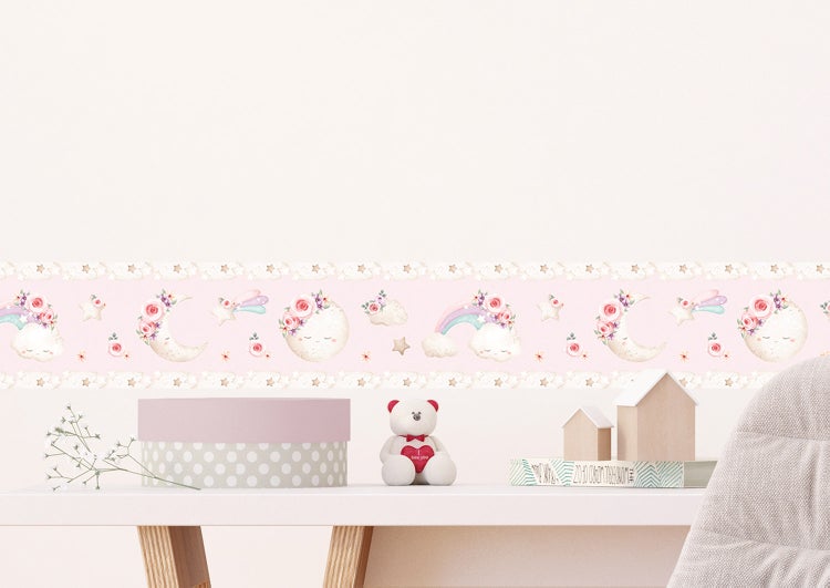 Hell gestaltete Kinderzimmerdekoration mit floraler Tapetenbordüre mit Mond- und Sternmotiven, Teddybär und Dekoelementen aus Holz auf einem Tisch.