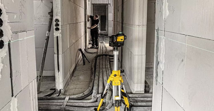 Ein Baustellenlaser auf einem Stativ projiziert eine horizontale Laserlinie in einem Rohbau während ein Arbeiter Messungen vornimmt.