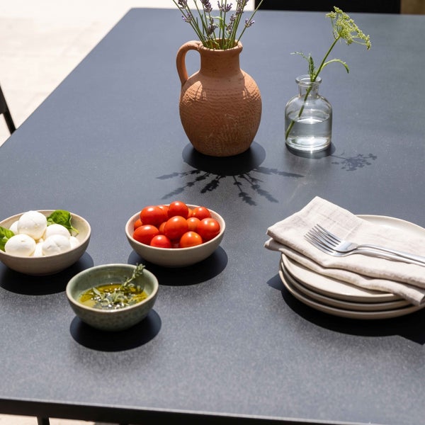 Moderner Gartentisch in Anthrazit mit Gedeck, Schüsseln mit Tomaten und Mozzarella, Terracotta-Krug und Glasvase bei hellem Tageslicht.