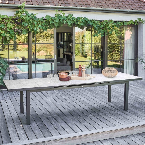 Ausziehbarer Gartentisch mit Platte in Steinoptik und dunklem Metallgestell auf einer Holzterrasse vor einer Glasfront.
