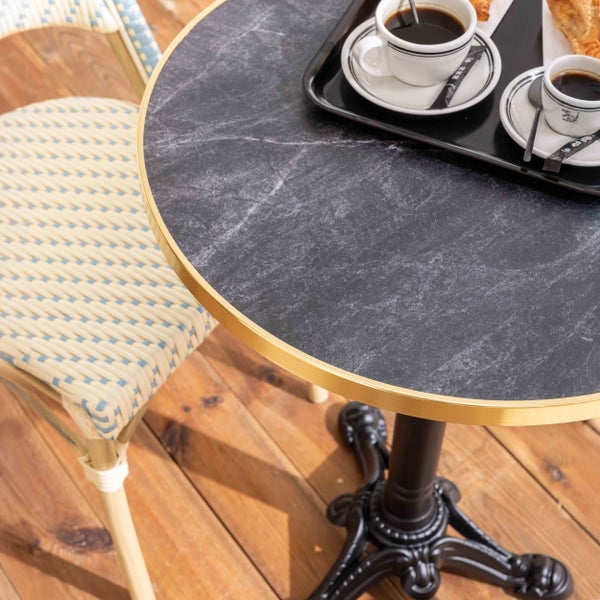 Runder Bistrotisch mit dunkler Marmor-Optik, goldfarbenem Rand und gusseisernem Fuß auf einer hellen Holzterrasse mit Kaffee und Gebäck.