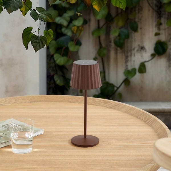 Braune Tischleuchte mit plissiertem Schirm auf einem runden Holztisch im Außenbereich, neben einem Glas Wasser.