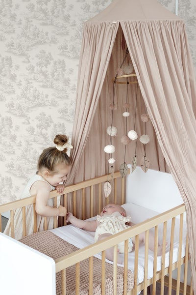 Helles Kinderzimmer mit Gitterbett aus Holz, Betthimmel aus Stoff, Mobile und Mustertapete. Ein Kleinkind steht am Bett bei einem Baby.