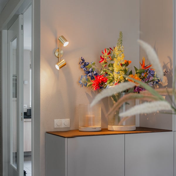 Wandleuchte mit zwei Spots neben einem Blumenarrangement auf einem Schrank