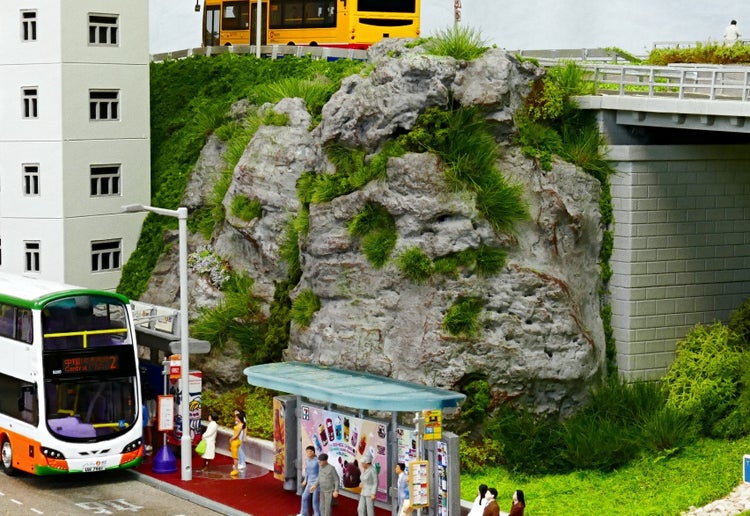 Modelleisenbahnlandschaft mit Felsen, Bushaltestelle, Doppeldeckerbus und Miniaturfiguren in einer städtischen Umgebung.