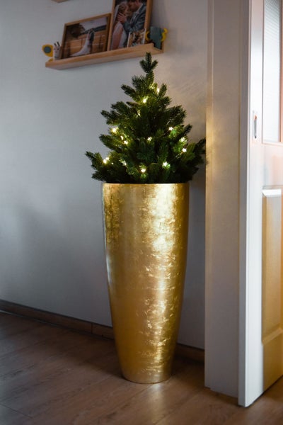 Kleiner künstlicher Weihnachtsbaum mit Beleuchtung in einer hohen goldfarbenen Bodenvase neben einer Tür.