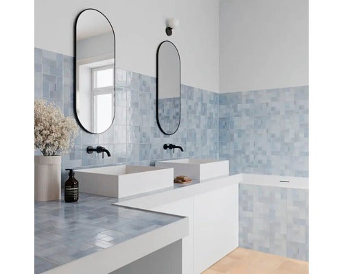 Modernes, helles Badezimmer mit hellblauen Wandfliesen, zwei weißen Aufsatzwaschbecken, schwarzen Armaturen und ovalen Spiegeln.