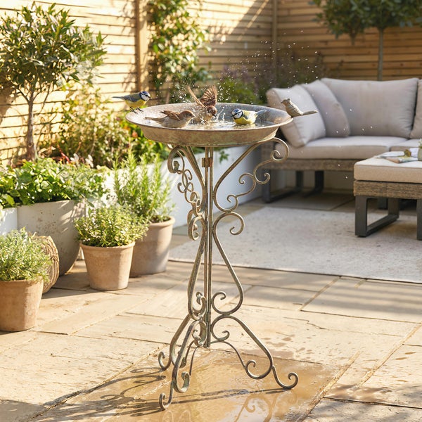 Dekorative Vogeltränke aus Metall mit verschnörkeltem Standfuß auf einer sonnigen Terrasse mit Vögeln und Gartenmöbeln.