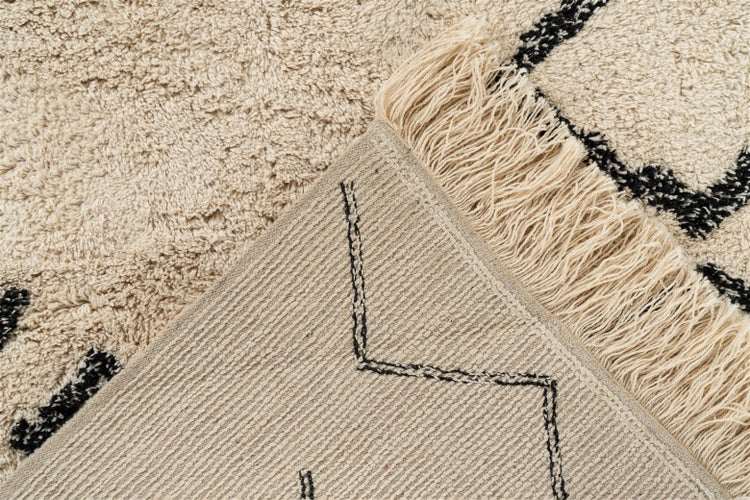 Hochflor-Teppich in Beige mit schwarzem geometrischem Muster, Fransen und einer umgeklappten Ecke, die die gewebte Rückseite zeigt.