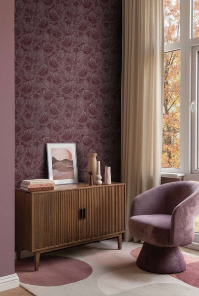 Wohnzimmer mit Vliestapete mit Blumenmuster, Holzkommode, gepolstertem Sessel und Fenster mit Ausblick auf herbstliche Bäume.