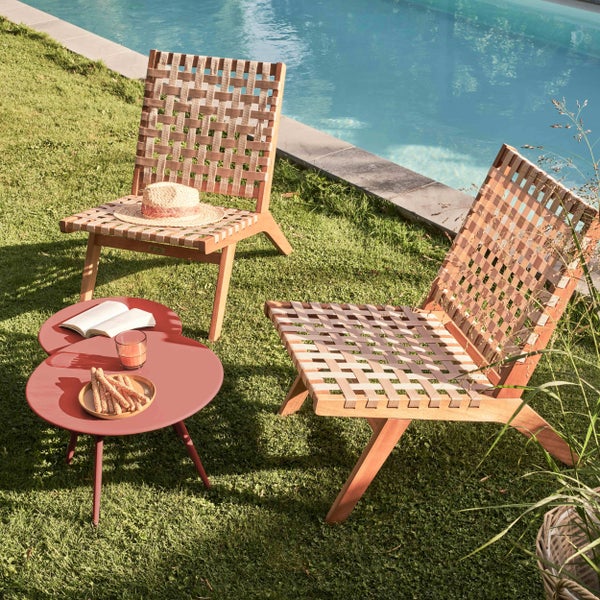 Zwei Garten-Loungesessel aus Holz mit Geflecht und ein roter Beistelltisch auf einer Rasenfläche neben einem Pool.