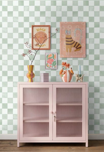 Vliestapete mit grün-weißem Karomuster hinter einem rosa Metallschrank mit Glastüren und bunter Dekoration.