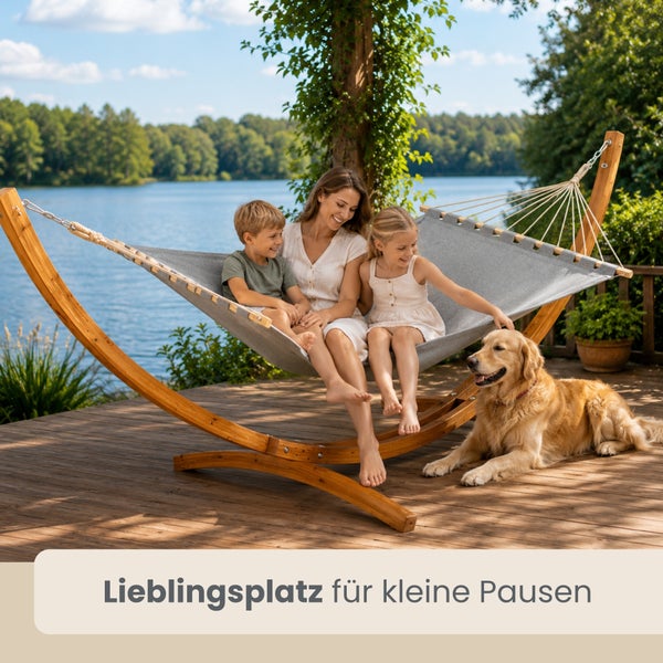 Graue Stoffhängematte mit Holzgestell auf einer hellen Holzterrasse am See mit einer Frau, zwei Kindern und einem Hund.