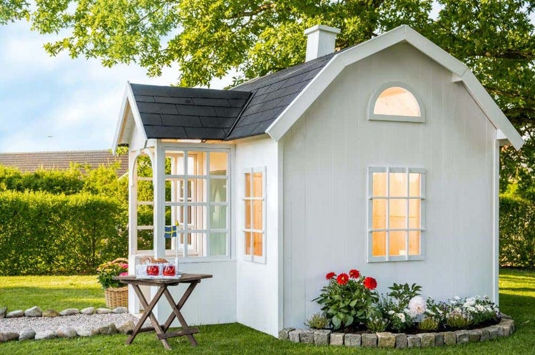 Weißes Kinderspielhaus aus Holz mit dunklem Satteldach, Sprossenfenstern und kleiner Veranda in einem Garten mit Blumenbeeten.