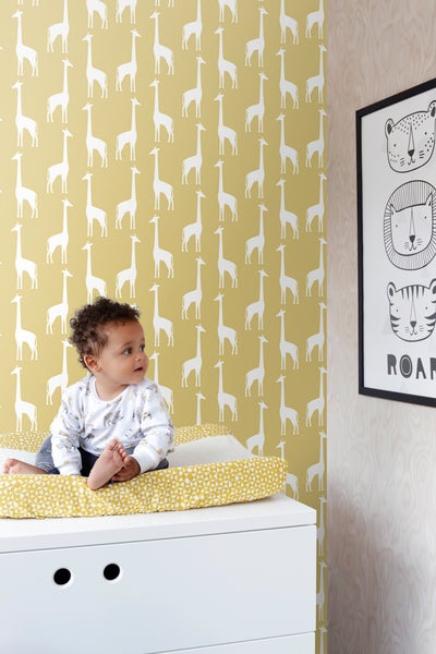 Ein Baby sitzt auf einer Wickelauflage auf einer weißen Kommode vor einer Tapete mit Giraffenmuster und einem gerahmten Bild mit Tiermotiven.
