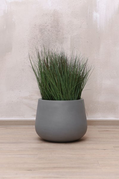 Künstliches Ziergras in einem runden grauen Pflanztopf auf einem Holzboden.