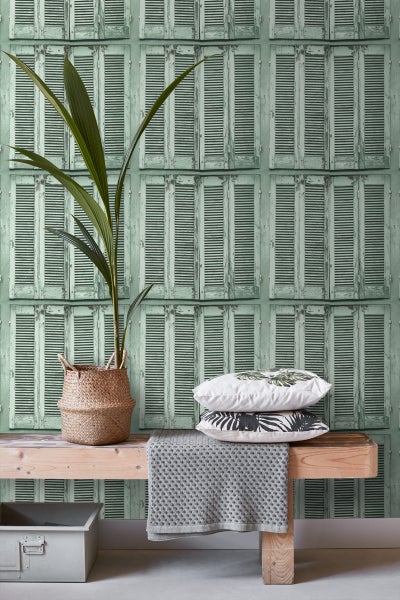 Szene mit einer Pflanze im Korb, Kissen und einer Bank vor einer Tapete mit Fensterladenmuster