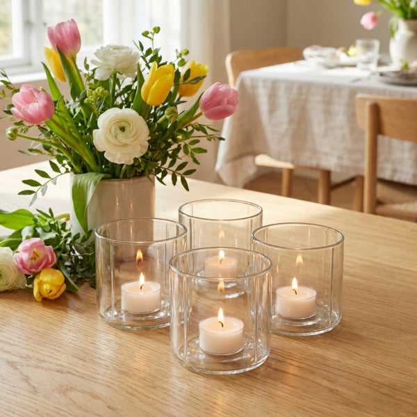 Vier zylindrische Teelichthalter aus Glas mit brennenden Teelichtern auf einem Holztisch neben einem Blumenstrauß