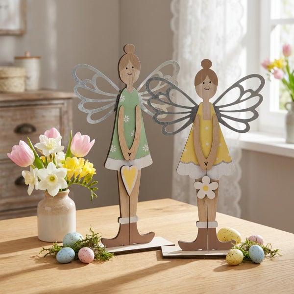 Zwei dekorative Holzfiguren als Osterfeen mit Metallflügeln auf einem Tisch mit Frühlingsblumen und Ostereiern als Osterdekoration.