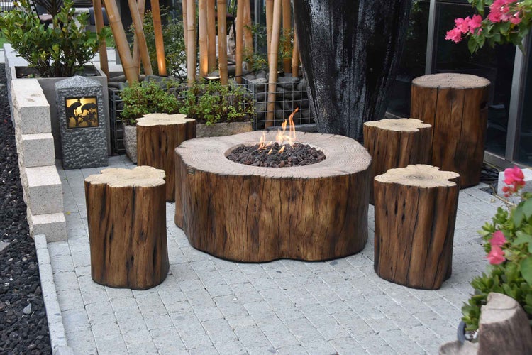 Rustikale Feuerstelle in Baumstamm-Optik mit passenden Hockern auf einer gepflasterten Terrasse im Garten.