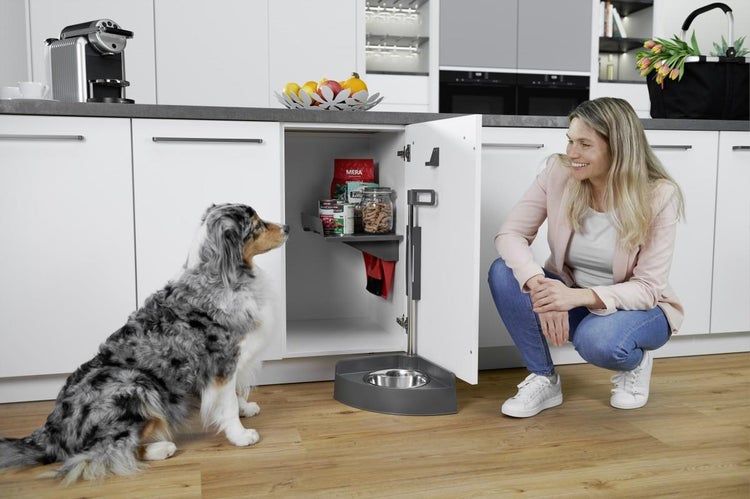 Moderne, helle Küche mit weißem Unterschrank und einer ausziehbaren Tierfutterstation. Eine Frau hockt neben einem Hund vor der geöffneten Tür.