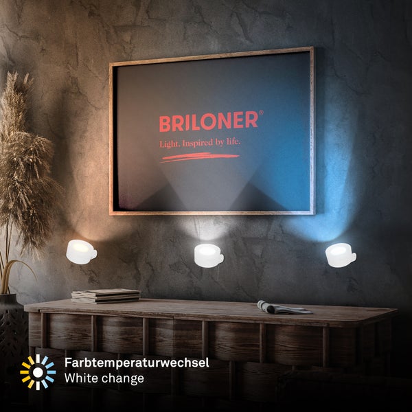 Drei LED-Spots mit einstellbarer Farbtemperatur von warmweiß bis kaltweiß an einer Wand über einer Holzkommode mit Briloner Logo.