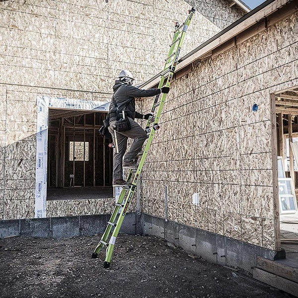 Handwerker mit Helm und Werkzeuggürtel auf einer grünen Mehrzweckleiter an einer Hauswand aus Holzwerkstoffplatten auf einer Baustelle.