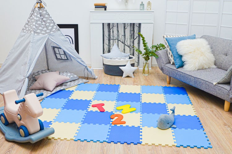 Modernes Kinderzimmer mit blau-beiger Schaumstoff-Puzzlematte, grauem Tipi-Zelt, Schaukelpferd und grauem Sofa auf hellem Holzboden.