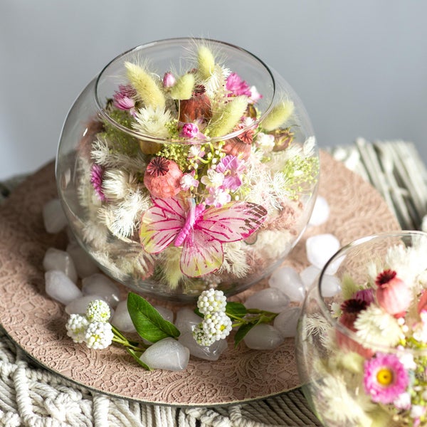 Dekorative Glaskugel mit einem Arrangement aus Trockenblumen und einem künstlichen Schmetterling, umgeben von dekorativen Steinen.