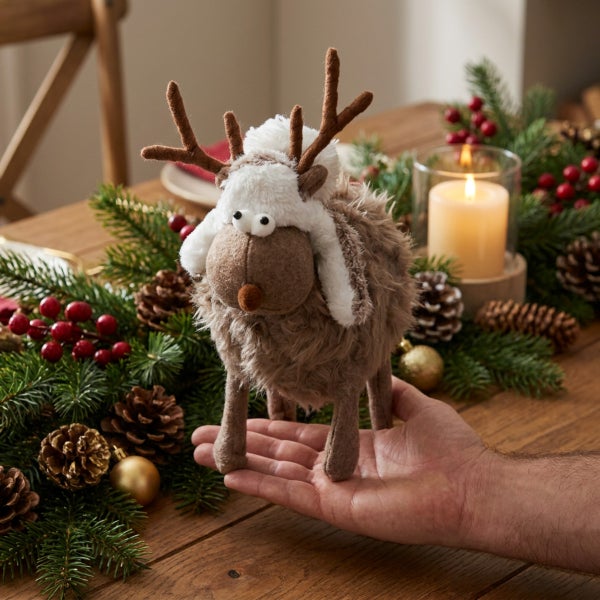 Eine Hand hält eine Plüsch Rentier Weihnachtsdekorationsfigur mit Felloptik und Ohrenschützer auf einem festlich dekorierten Holztisch.