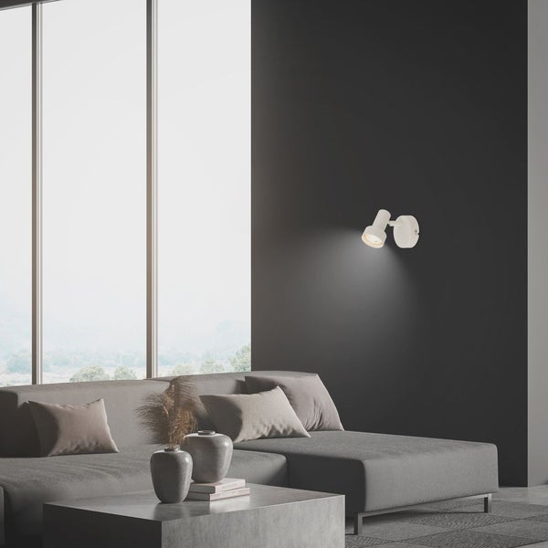 Moderne Wandleuchte in einem hellen Wohnzimmer mit grauem Ecksofa, Betontisch und großen Fenstern.